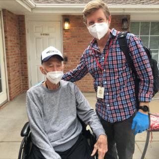 Two individuals wearing face masks are outside, one seated in a wheelchair and the other standing beside them.