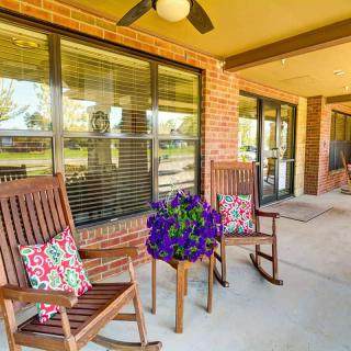 A cozy porch featuring two wooden rocking chairs with colorful cushions, a small table adorned with vibrant flowers, and large windows set into a brick wall.