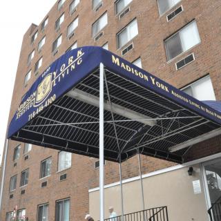 The entrance of a hotel with a blue awning is seen against a brick building backdrop.