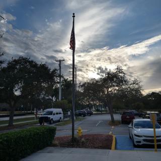 A flagpole stands tall in a parking lot with cars and trees under a cloudy sky.