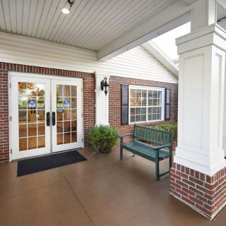 A welcoming building entrance with double glass doors, a bench, and decorative plants.