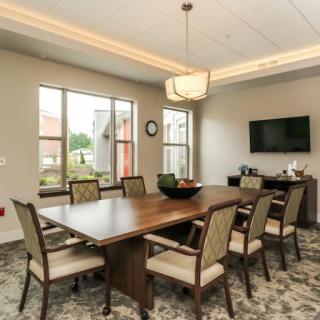A modern conference room with a large table, several chairs, windows, and a wall-mounted television.