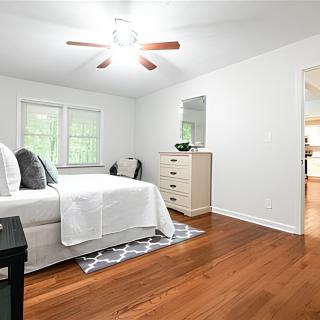 A bright and spacious bedroom with hardwood floors, featuring a neatly made bed, a dresser, and a ceiling fan.
