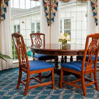 A cozy dining room with a wooden table and chairs, featuring a glass tabletop and blue cushioned seats. Large windows with patterned curtains adorn the space, accompanied by a decorative carpet and an indoor plant.
