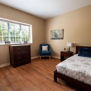 A cozy bedroom with wooden flooring, featuring a bed, a dresser, and a chair next to a window.