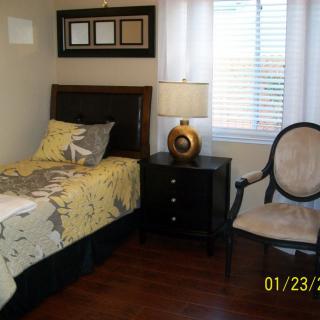 A cozy bedroom with a single bed, nightstand, lamp, and chair by the window.