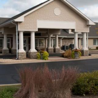 A single-story building with prominent columns at the entrance, surrounded by neatly maintained landscaping and a paved parking area under a cloudy sky.