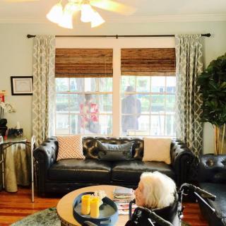 A cozy living room with a black leather sofa, large windows with curtains, and a ceiling fan above.