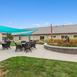 A sunny outdoor patio area with tables and chairs under a shade structure, surrounded by landscaped gardens.