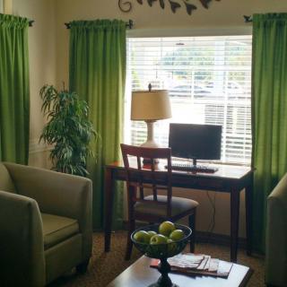 A cozy living room setup with green curtains, featuring a desk with a computer and lamp by the window.