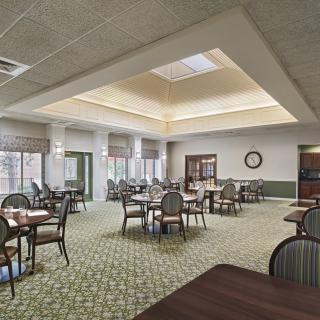 A spacious dining room with multiple tables and chairs, featuring carpeted flooring and large windows that let in natural light.