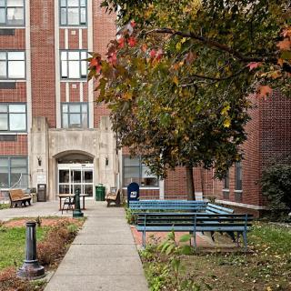 The entrance of a brick building surrounded by trees and benches, featuring a well-maintained pathway.