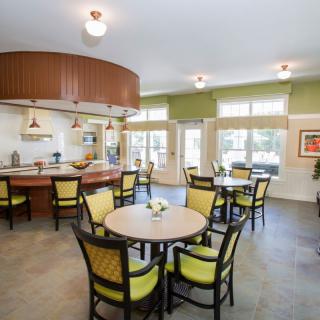 A spacious and well-lit dining area with round tables and cushioned chairs near an open kitchen.