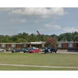 A school building with a parking lot filled with cars, an American flag flying, and trees in the background.