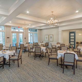 A spacious and elegantly decorated dining room with multiple tables and chairs, featuring a chandelier and large windows.