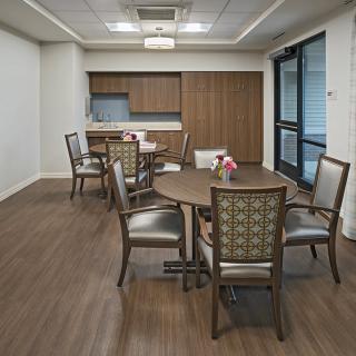 A modern dining room with a wooden floor, featuring tables and chairs, adjacent to a kitchen area.