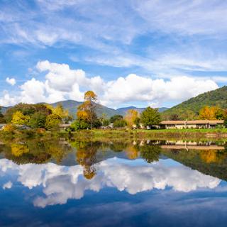 A serene view of a lake reflecting the colorful autumn trees and vibrant clouds against the backdrop of rolling mountains.