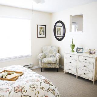 A bright and cozy bedroom with neutral tones, featuring an armchair, a dresser, and decorative artwork.