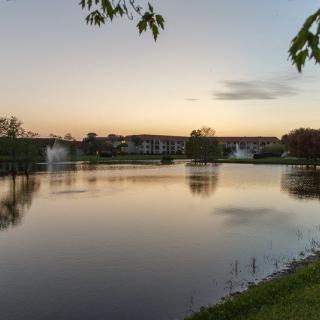 A tranquil lakeside view at sunset with calm waters reflecting the colorful sky and nearby residential buildings.