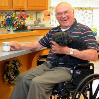 An elderly man sitting in a wheelchair, holding a dog and smiling in a kitchen.