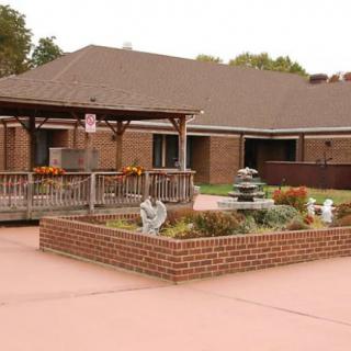 A brick building with a garden and fountain in the foreground, alongside a wooden gazebo.
