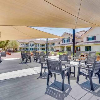 A shaded outdoor patio area with tables and chairs, adjacent to a large building on a sunny day.