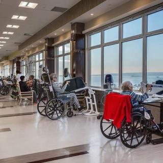 Elderly residents in a nursing home sitting by large windows enjoying the sunlight.