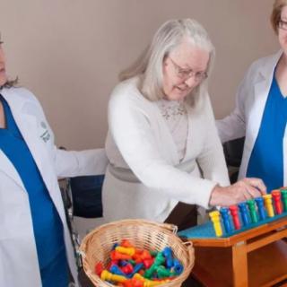 Healthcare workers assist an elderly woman in a therapeutic activity involving a xylophone.