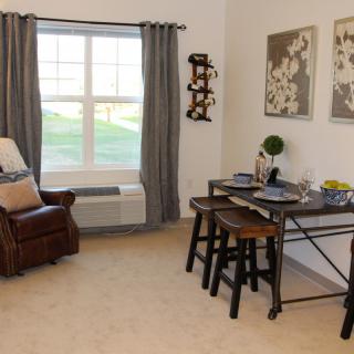 A cozy living and dining area with a large window, featuring comfortable seating, decorative artwork, and a neatly set table.