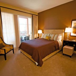 A cozy bedroom with warm tones, featuring a bed, nightstands with lamps, and a chair near a window.
