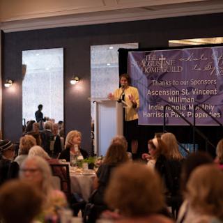 A speaker presents at a conference, engaging an attentive audience seated at tables.