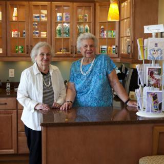 Two elderly women standing in a kitchen next to a display of greeting cards, smiling at the camera.