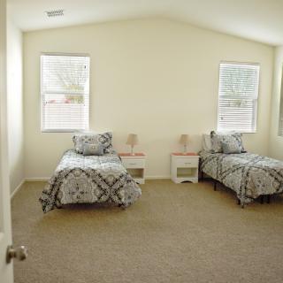 A minimalist bedroom featuring twin beds with patterned bedding, carpeted flooring, and two windows providing natural light.