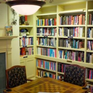 A cozy library room with a large bookshelf, a table with chairs, and a fireplace.