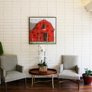 A cozy seating area featuring a red barn painting, two comfortable chairs, and a round table adorned with greenery.