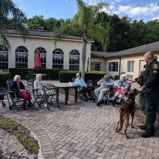 A group of elderly residents enjoys a visit from a police officer and his dog on the patio of a nursing home.