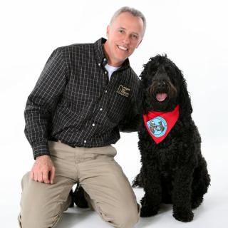 A man is kneeling next to a black dog wearing a red bandana.