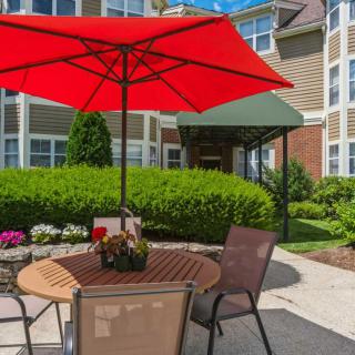 A cozy outdoor patio with a red umbrella, surrounded by lush greenery and flowers.