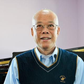 An elderly man with glasses wearing a blue vest and shirt indoors.