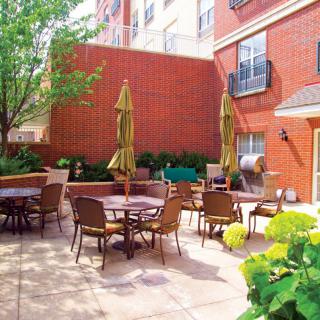 A cozy outdoor patio area with multiple tables and chairs, shaded by umbrellas, surrounded by a brick building and greenery.