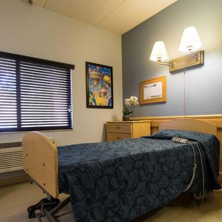 A hospital room with a single bed, wall art, and window blinds.