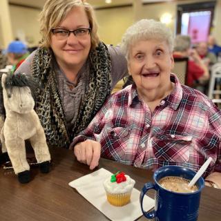 An elderly woman enjoying a cupcake and coffee, smiling alongside a companion indoors.