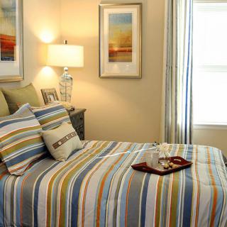 A cozy bedroom with striped bedding, decorative pillows, framed wall art, and a table lamp.