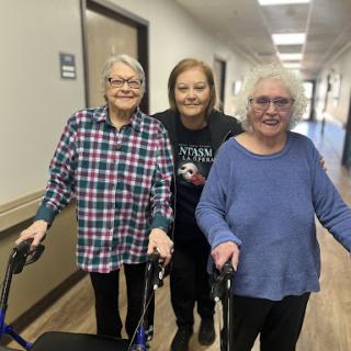Two elderly women assisted by a caregiver as they walk down the hallway.