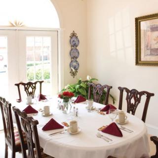 A bright dining room with a round table set for a meal, featuring red napkins and a centerpiece of flowers.