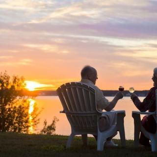 A couple enjoys a peaceful sunset by the lake, toasting with glasses while seated in outdoor chairs.