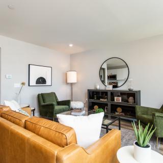 A modern living room with a brown leather sofa, green chairs, and a round mirror above a shelf unit.