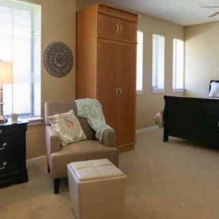 A cozy bedroom featuring a chair with cushions, a lamp on a nightstand, and several windows allowing natural light.