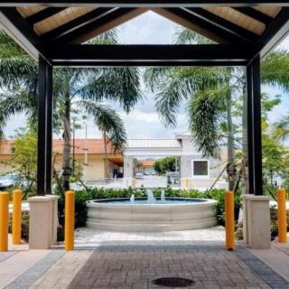 A view through a covered walkway towards a fountain surrounded by palm trees in an urban setting.
