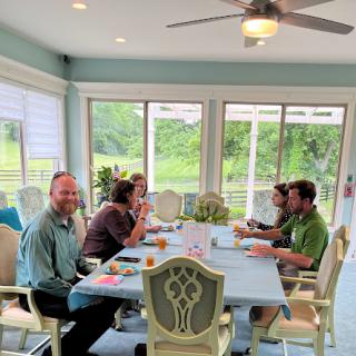 A group of friends gathered around a table in a sunroom, enjoying breakfast and engaging in conversation.